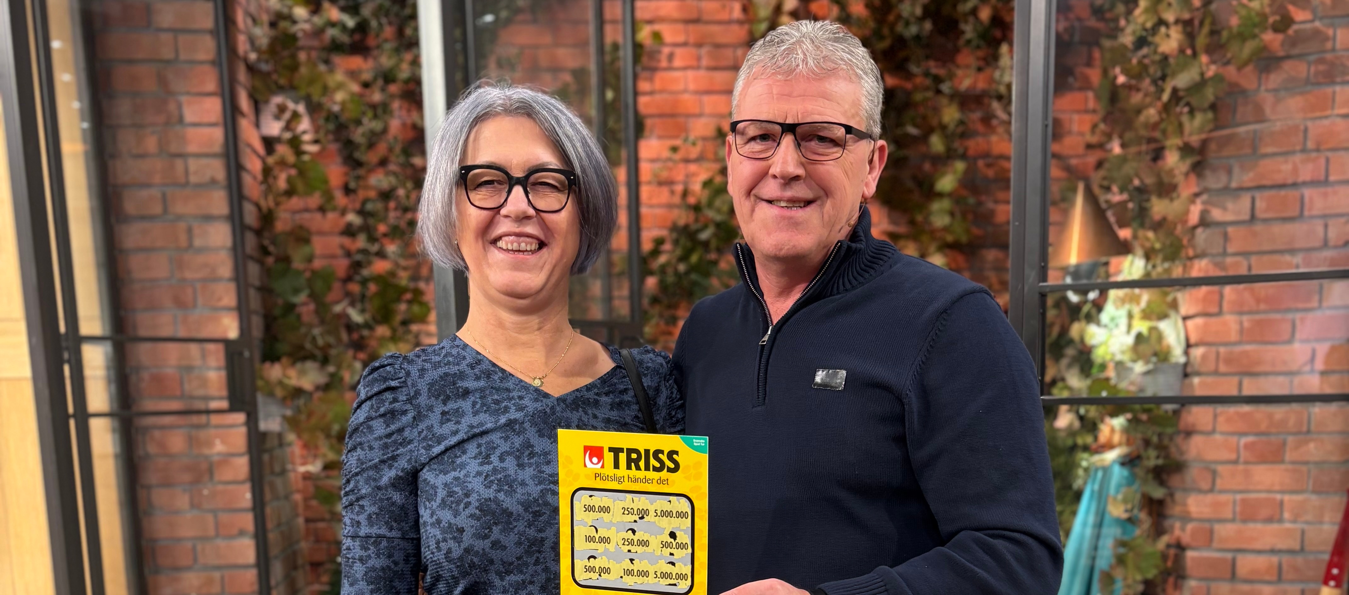 Två glada personer poserar med en gul Triss-lott framför en vägg av tegel och grönt lövverk.