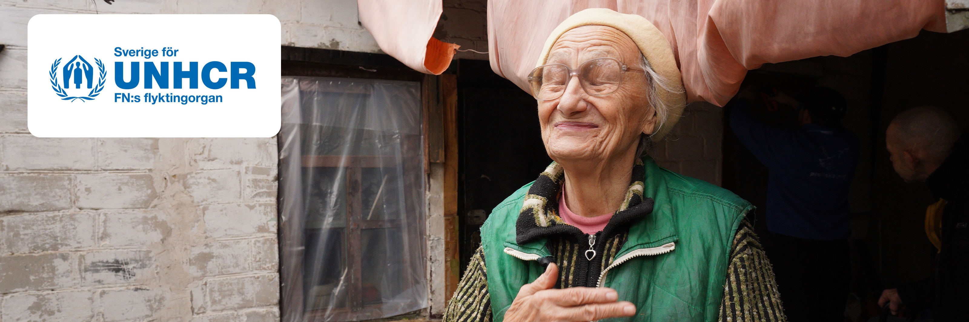 Äldre kvinna i grönt står utomhus vid hus, under Sverige för UNHCR-logotyp.