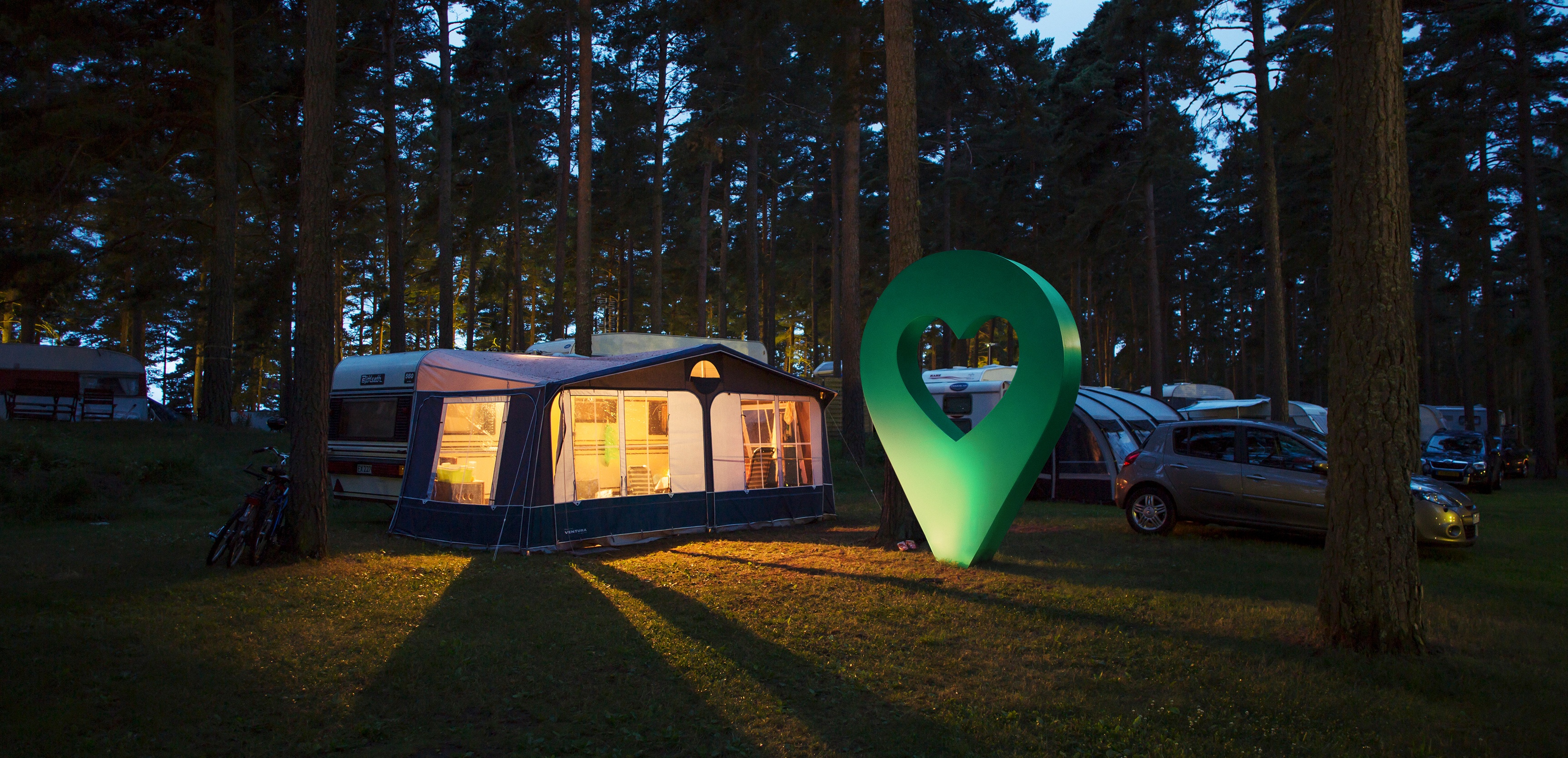 Kvällsbild på en tältplats i en skog med en stor grön markör som lyser. Campingtältet är upplyst inifrån.
