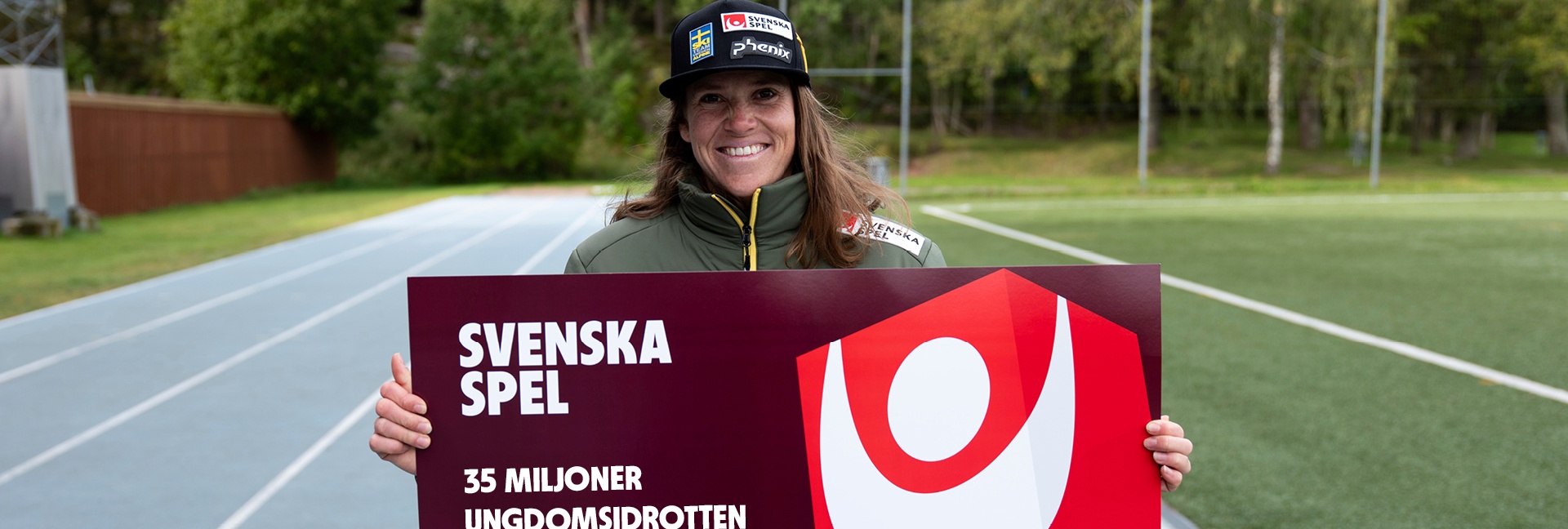 Sara Hector håller en skylt på en löparbana med text om Svenska Spel och ungdomsidrotten Gräsroten.